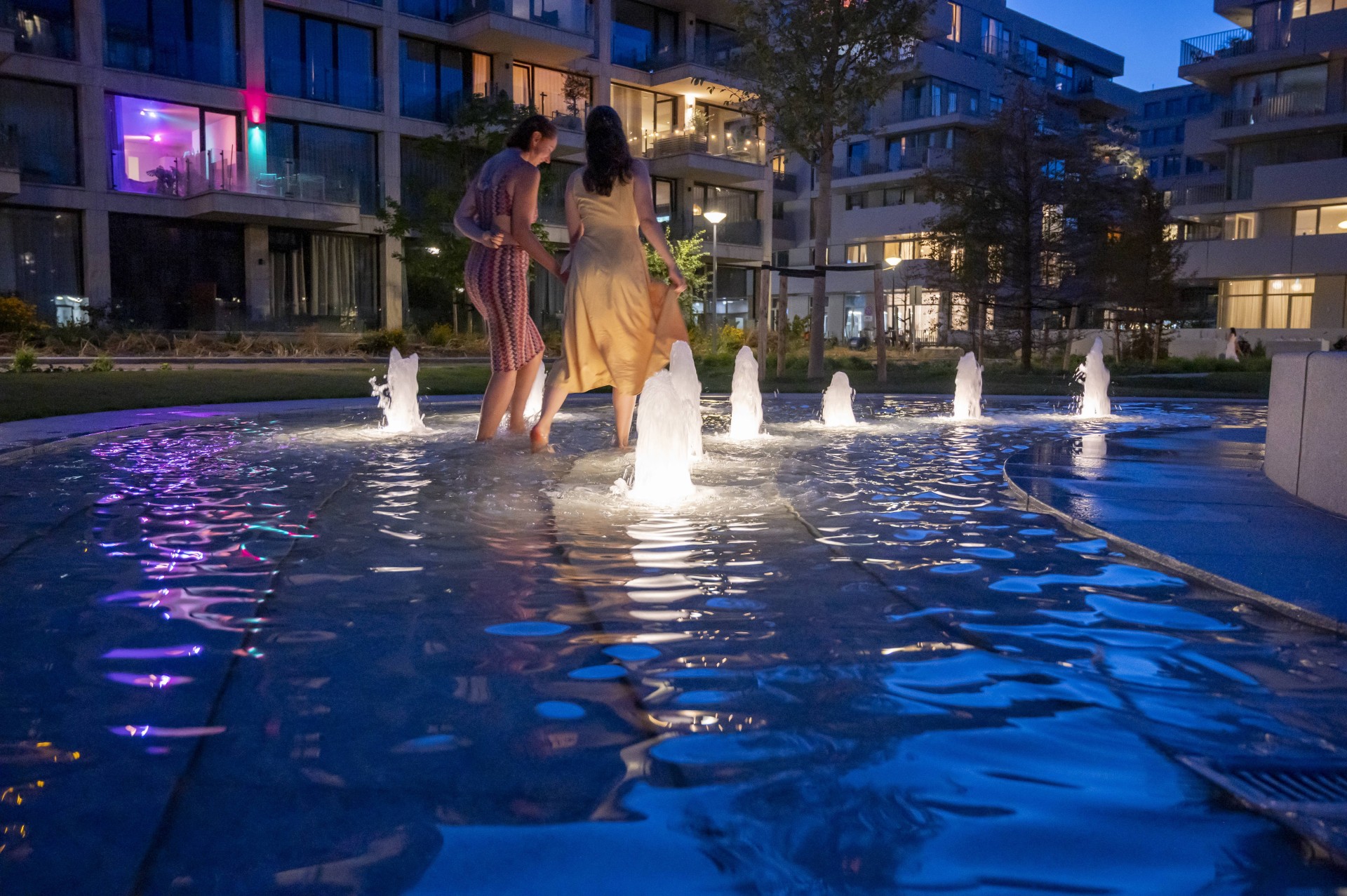 Kinderen bij de verlichte spuiters van de fontein in de avond op het Buurtplein