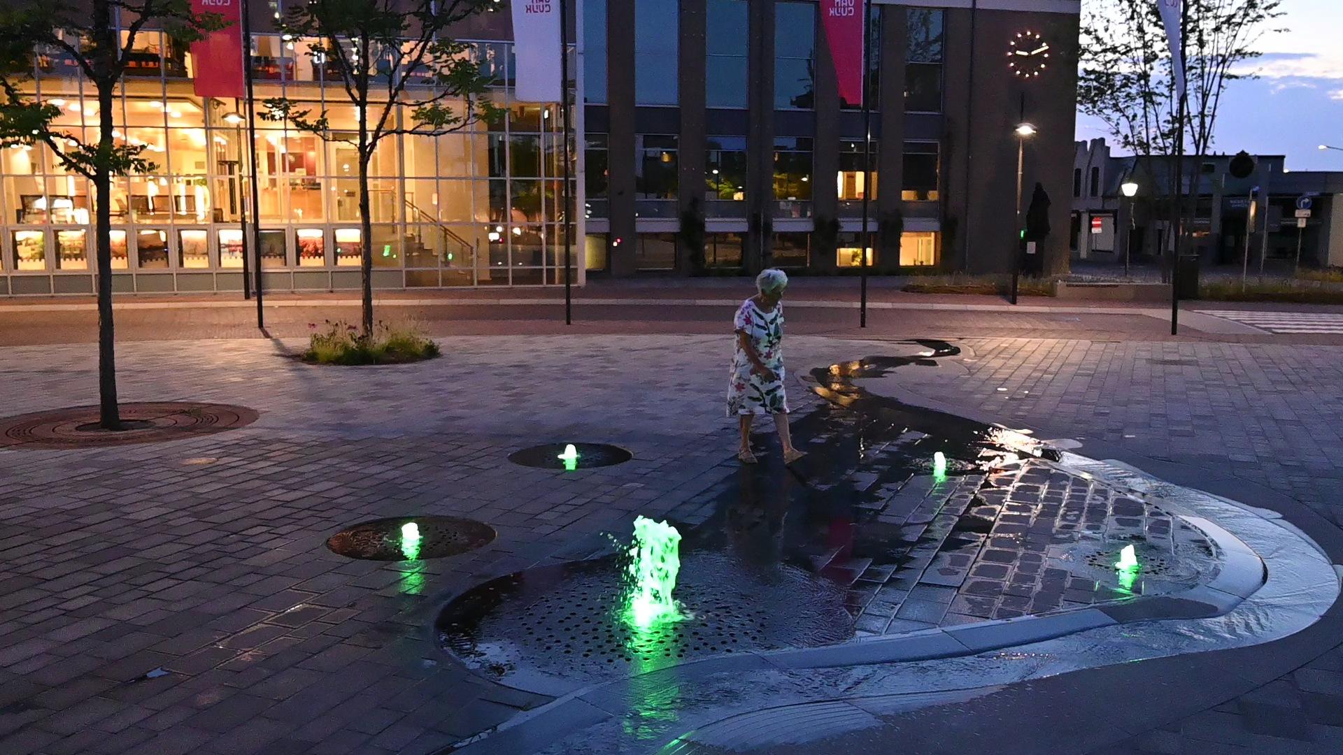 Bezoeker loopt 's avonds door het water van de groen verlichte fonteinen op het plein in Cuijk