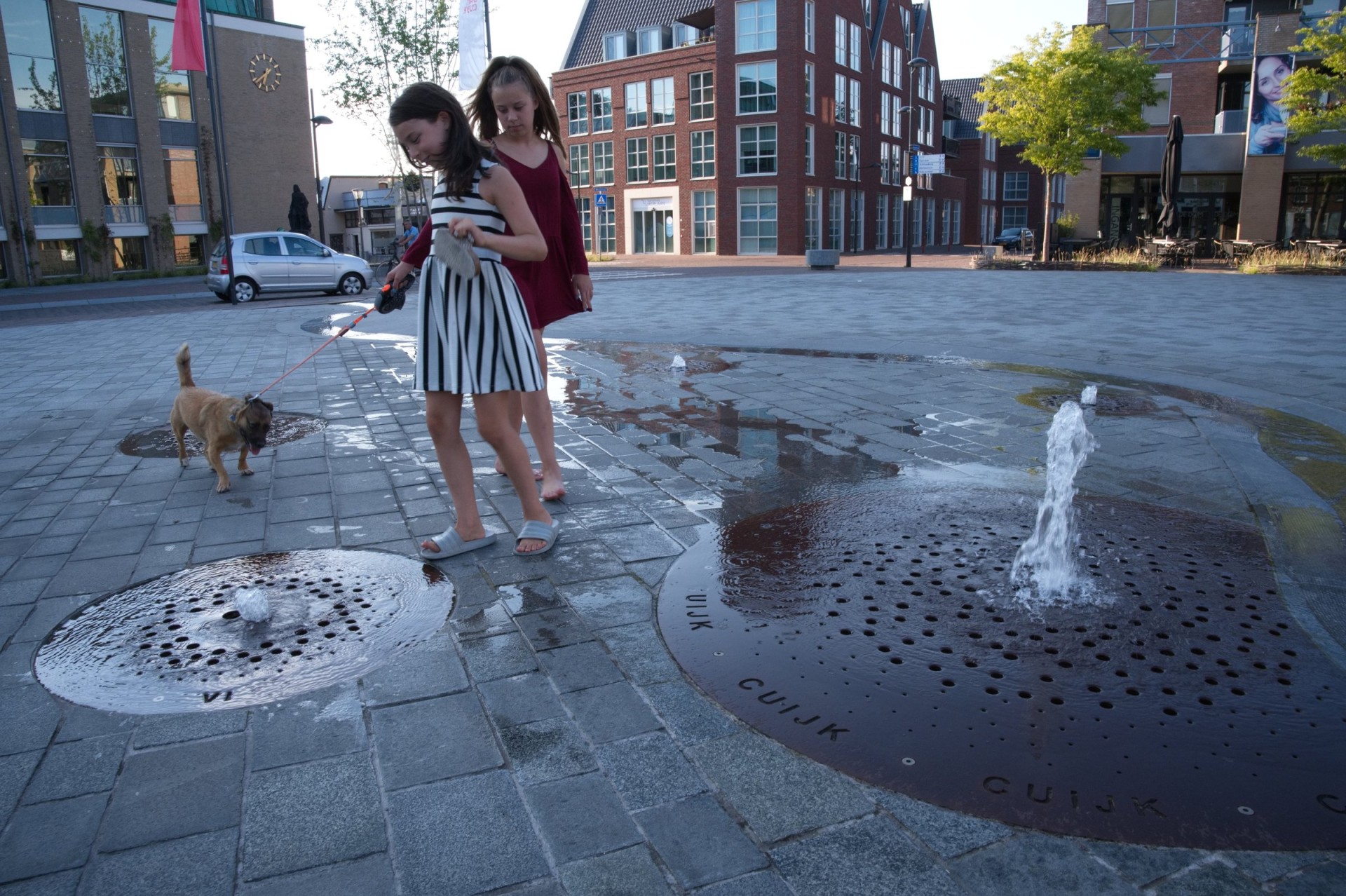 Cuijk meisjes hond spelen fontein spuiter cortenstaal plein Louis Jansenplein 25 Meisjes spelen met een hond bij de cortenstalen spuiters op het Louis Jansenplein in Cuijk
