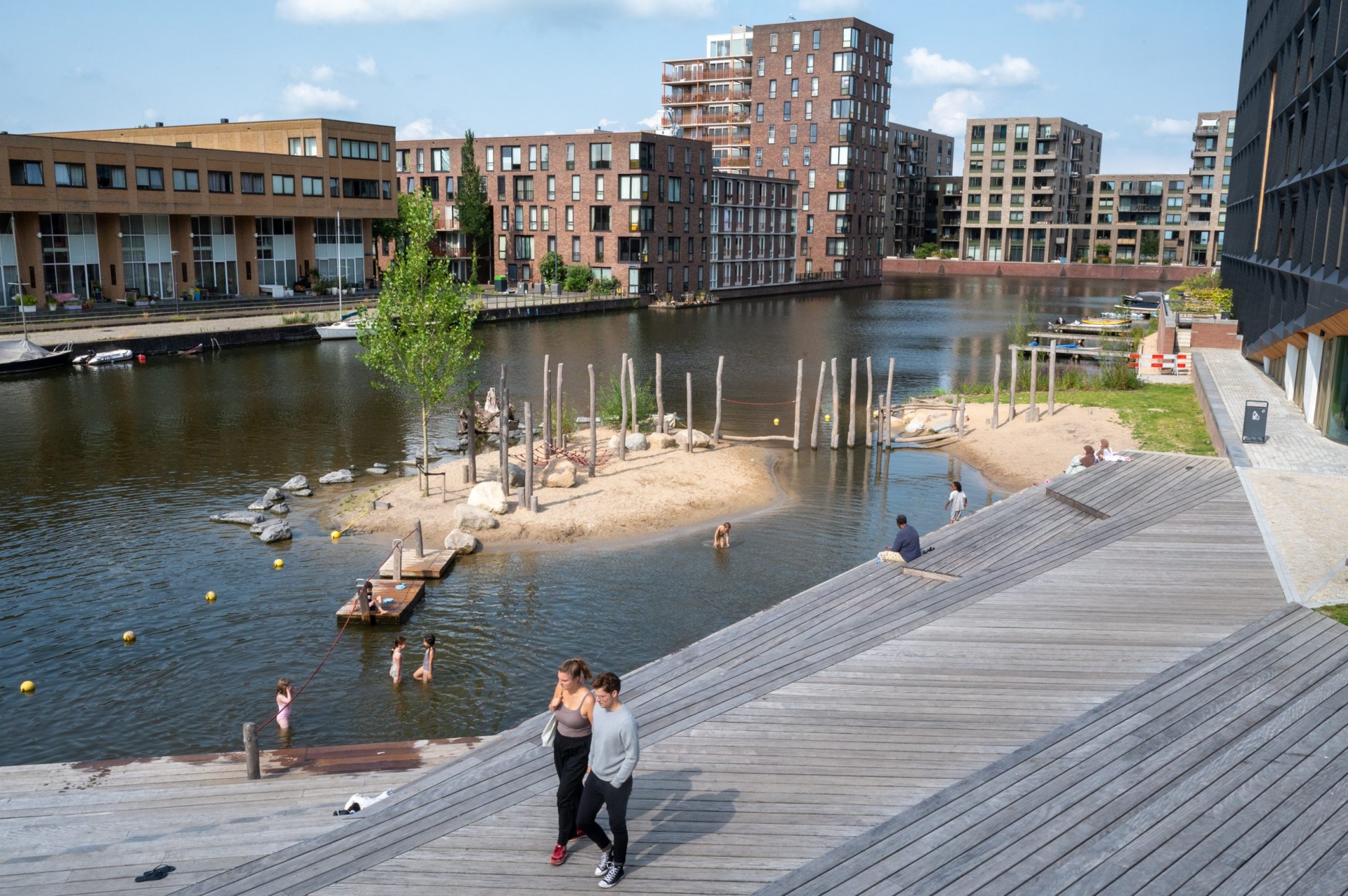Wandelaars op de houten boardwalk langs het zandeiland in de haven bij Jonas in IJburg
