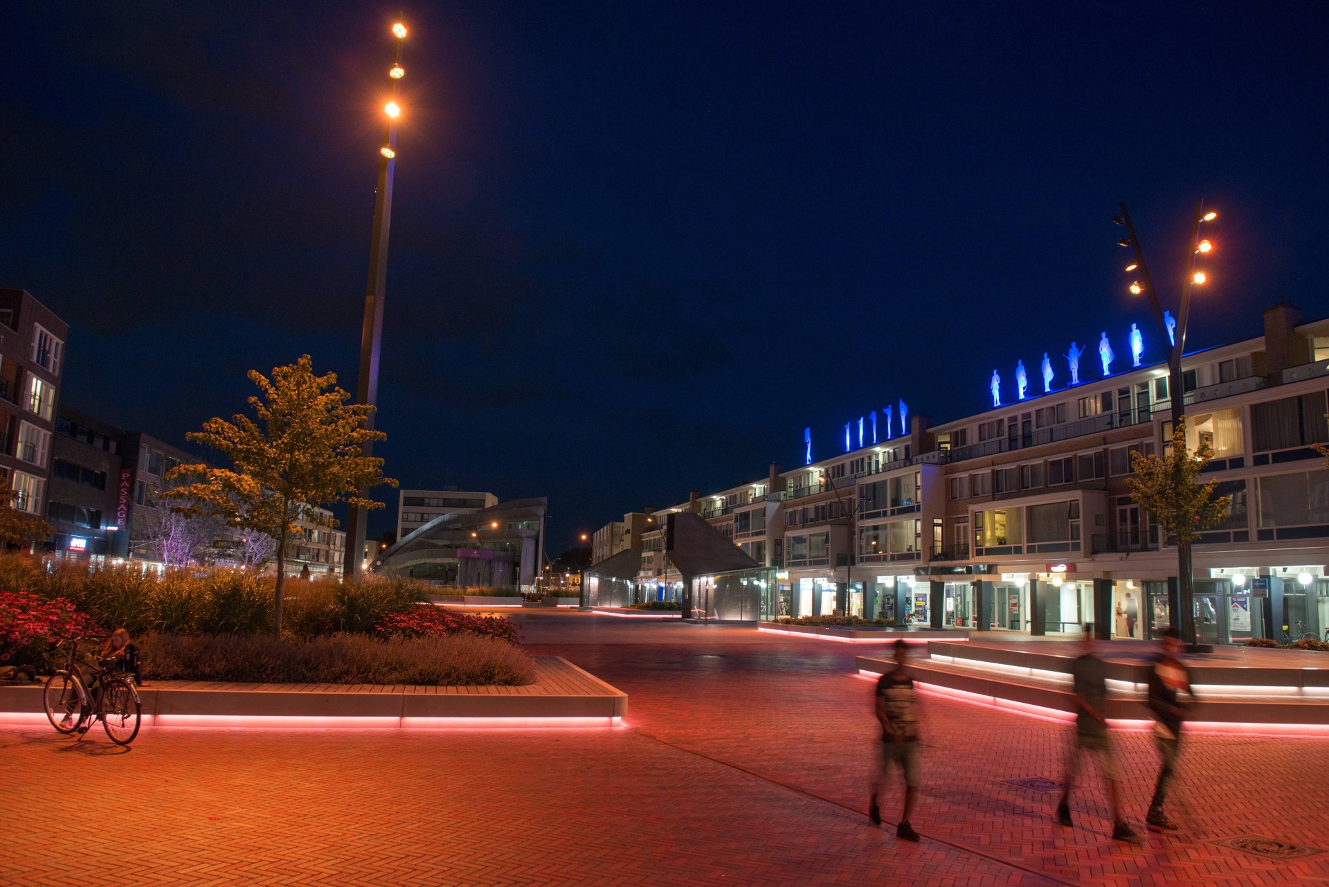Nieuwe Markt Roosendaal wandelaars avond LED lichtlijn betonband groen eiland 111 Wandelaars langs een LED-lichtlijn in een betonband bij een groen eiland op de Nieuwe Markt in Roosendaal