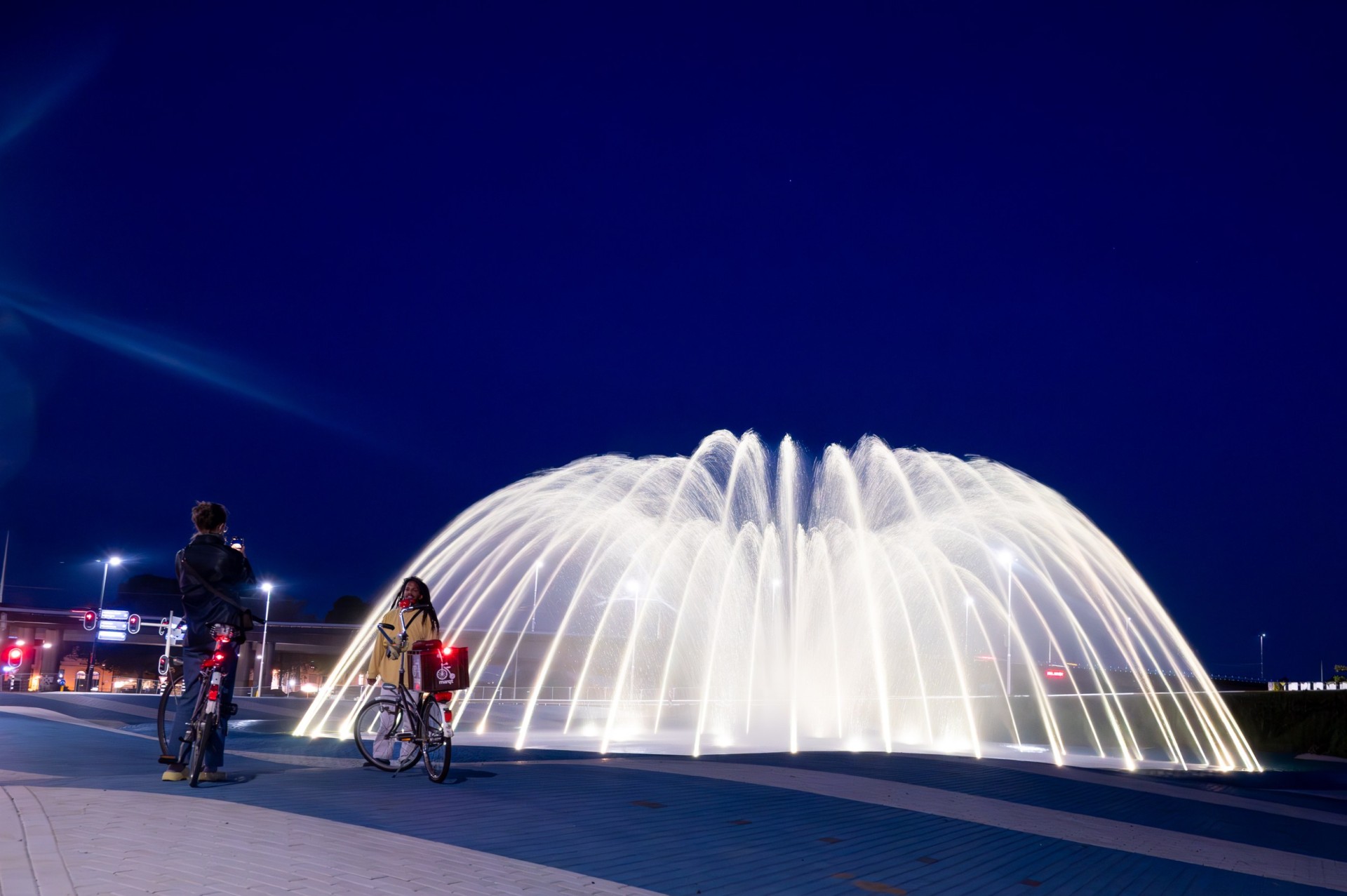Roermondsplein: lichtgevende waterfontein in de avond met fietsers op de voorgrond